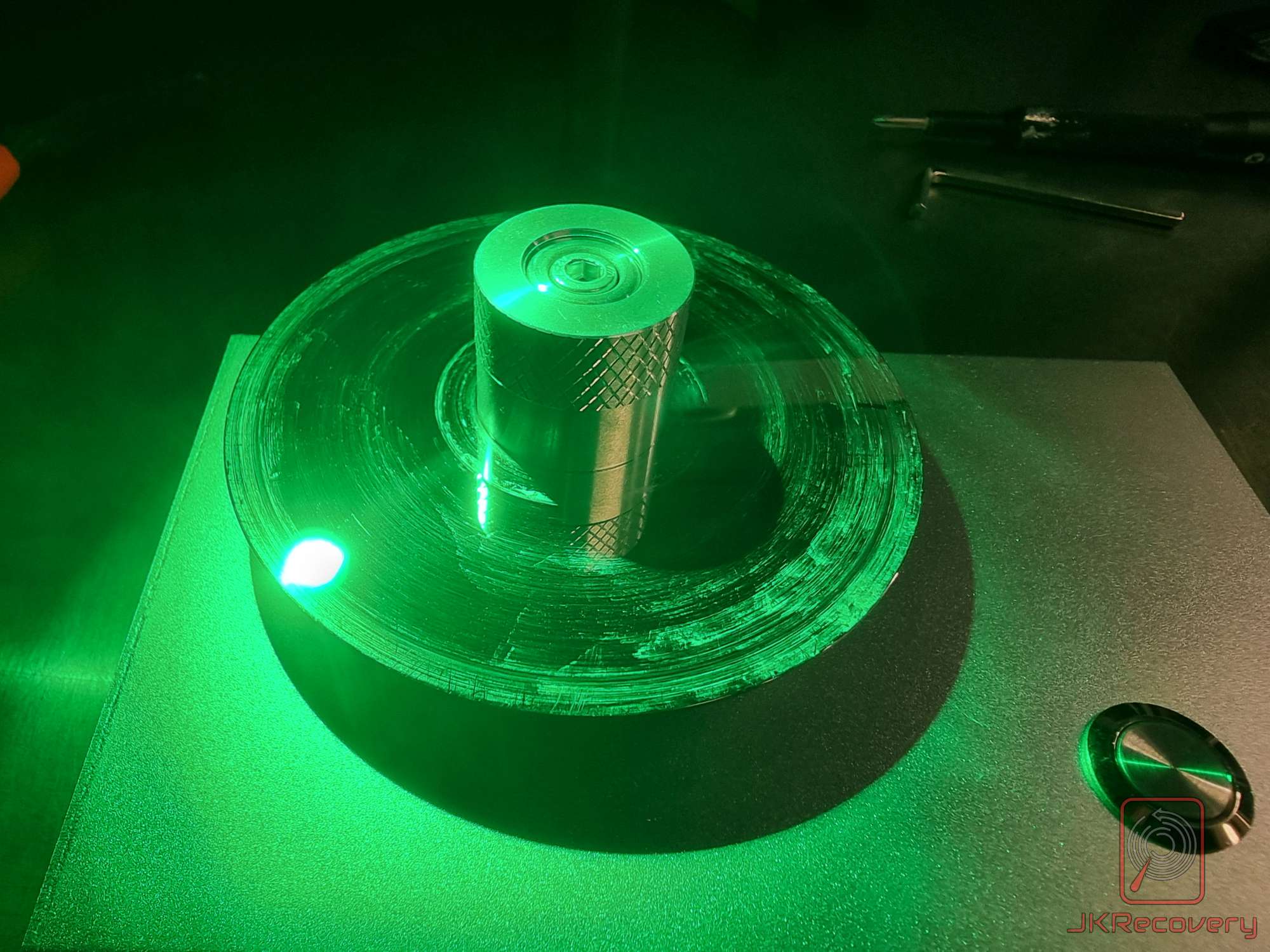 Talerz dysku twardego HDD oświetlony zielonym światłem UV w laboratorium odzysku danych, widoczny uchwyt do mocowania talerza oraz śruba centrująca